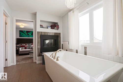 Bathroom featuring a freestanding bathtub with brushed nickel fixtures, a wall-mounted fireplace with a decorative tiled surround, and large windows with white curtains - 18 Enchanted Way, St. Albert, AB 
