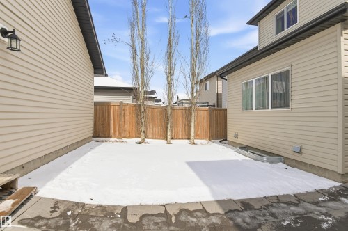 The property features beige siding, a partial concrete patio, and a fenced yard with three tall, bare trees - 678 Secord Boulevard, Edmonton, AB - Outdoor With Exterior