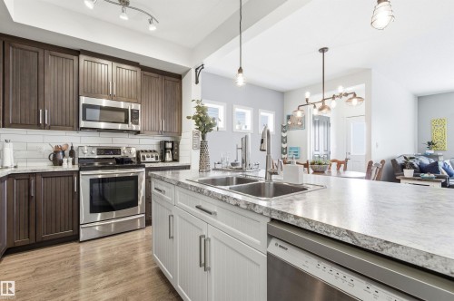The kitchen features dark wood cabinetry, stainless steel appliances, a white subway tile backsplash, and a large island with a double sink and light-colored countertop - 678 Secord Boulevard, Edmonton, AB - Indoor Photo Showing Kitchen With Double Sink With Upgraded Kitchen