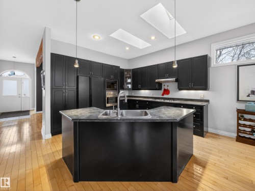 The kitchen features a central island with a double sink, dark cabinetry, stainless steel appliances, and light wood flooring - 569 Butterworth Way, Edmonton, AB - Indoor Photo Showing Kitchen With Double Sink With Upgraded Kitchen
