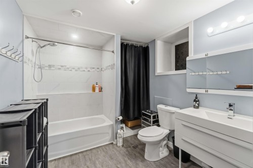 Bathroom featuring a white bathtub with subway tile surround and decorative accent band, a built-in shower niche, a white vanity with integrated sink and chrome faucet, a wall-mounted mirrored cabinet, and wood-finish flooring - 6036 106 Street, Edmonton, AB - Indoor Photo Showing Bathroom