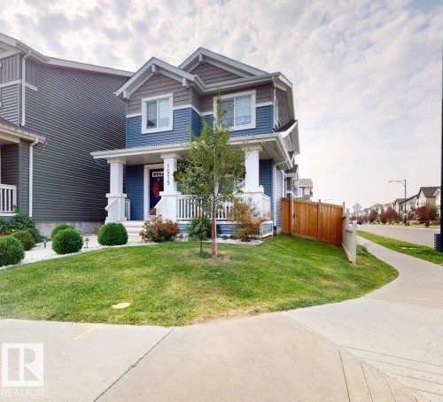 The property features a blue and gray exterior with white trim, a covered front porch with white railings, and a well-maintained lawn with a young tree and shrubbery - 1275 Chappelle Boulevard, Edmonton, AB 