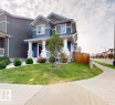 The property features a blue and gray exterior with white trim, a covered front porch with white railings, and a well-maintained lawn with a young tree and shrubbery - 1275 Chappelle Boulevard, Edmonton, AB 
