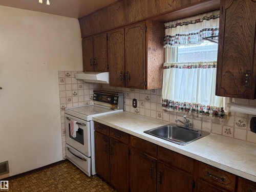 The kitchen features wood cabinetry, a white stove with an overhead exhaust fan, a double basin stainless steel sink, and light-colored countertops - 10916 151 Street, Edmonton, AB - Indoor Photo Showing Kitchen