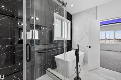 Luxurious bathroom featuring a spacious glass-enclosed shower with black marble-style tiling, a free-standing white soaking tub with a black floor-mounted faucet, and white marble-style wall and floor tiling - 190 Elliott Wynd, Fort Saskatchewan, AB - Indoor Photo Showing Other Room