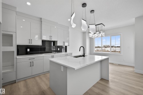 The kitchen features light-toned cabinetry with dark handles, a dark tiled backsplash, and a central island with a white countertop and an integrated sink with a black faucet - 190 Elliott Wynd, Fort Saskatchewan, AB - Indoor Photo Showing Kitchen With Double Sink With Upgraded Kitchen