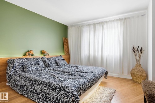 This bedroom features warm-toned wooden flooring, a light green accent wall, and a large window dressed with white sheer curtains - 11026 110 Avenue, Edmonton, AB - Indoor Photo Showing Bedroom