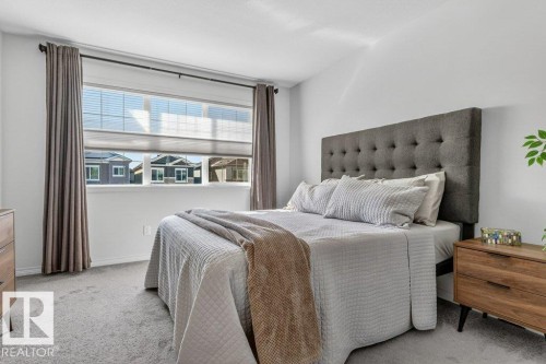 The bedroom features a large window with blinds and drapes, light gray carpet, and a tufted upholstered headboard - 3708 42 Avenue, Beaumont, AB - Indoor Photo Showing Bedroom