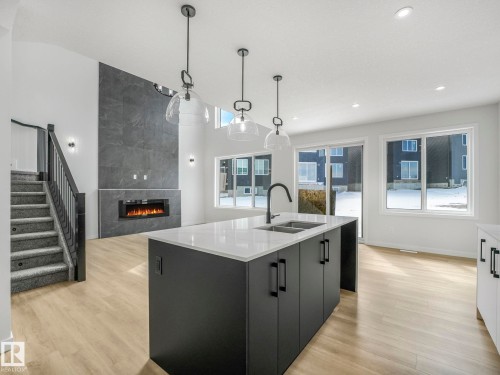Open concept living space featuring light-toned flooring, a kitchen island with a double sink and dark cabinetry, and an electric fireplace set into a dark tile wall - 239 Kinglet Boulevard Nw, Edmonton, AB - Indoor Photo Showing Kitchen With Double Sink With Upgraded Kitchen