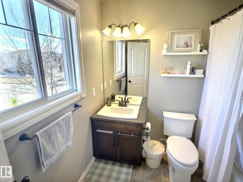 The bathroom features a window, a vanity with a sink and storage, a toilet, and a shower with a white curtain - 5718 Rue Eaglemont, Beaumont, AB - Indoor Photo Showing Bathroom