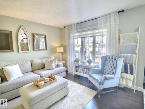 Living room featuring light-colored walls, dark flooring, and a large window with sheer curtains - 5718 Rue Eaglemont, Beaumont, AB - Indoor Photo Showing Living Room