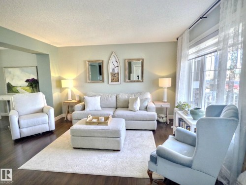 This inviting living area features dark wood flooring, light-colored walls, and a large window with sheer white curtains - 5718 Rue Eaglemont, Beaumont, AB - Indoor Photo Showing Living Room