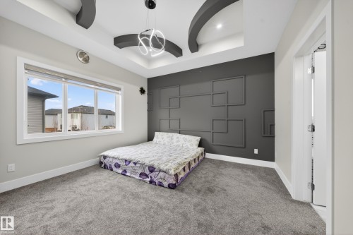 This room features a decorative accent wall with geometric patterns, a tray ceiling with integrated lighting and a modern light fixture, and a large window providing natural light - 449 Roberts Crescent, Leduc, AB - Indoor Photo Showing Bedroom
