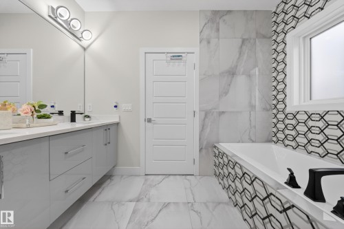 The bathroom features a double vanity with grey cabinetry and a white countertop, a bathtub with black and white hexagonal tile surround, and a large window providing natural light - 449 Roberts Crescent, Leduc, AB - Indoor Photo Showing Bathroom