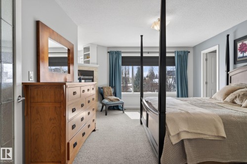 This bedroom features neutral carpeting, a large window with blue curtains, and a built-in fireplace with shelving - 20719 51 Avenue, Edmonton, AB - Indoor Photo Showing Bedroom