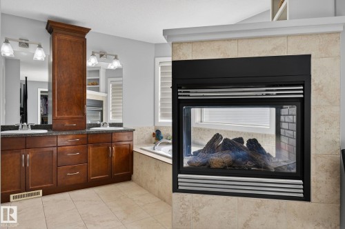 Bathroom featuring a double vanity with dark wood cabinetry, dual mirrors with sconce lighting, and a tiled fireplace with an insert - 20719 51 Avenue, Edmonton, AB - Indoor With Fireplace