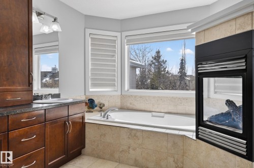 This bathroom features a built-in bathtub, large windows with horizontal blinds, a fireplace with a stone surround, and dark wood cabinetry with a sink and countertop - 20719 51 Avenue, Edmonton, AB - Indoor Photo Showing Bathroom