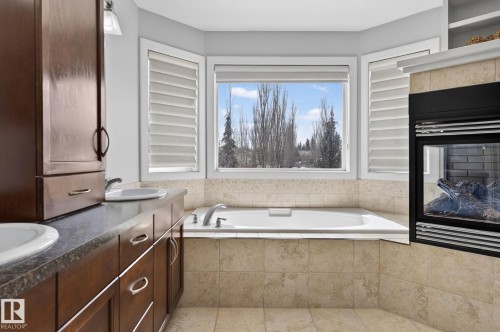 Bathroom featuring an in-built bathtub with tiled surround, a large window providing natural light, a fireplace, and a double vanity with two sinks - 20719 51 Avenue, Edmonton, AB - Indoor Photo Showing Bathroom