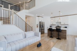 The property features an open-concept living area with light-toned flooring, a staircase with light wood and black metal railings, and a kitchen with white cabinetry and a stainless steel refrigerator - 
