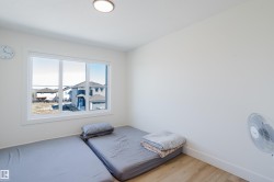 The property features a room with light wood-tone flooring, a large window, white walls, and a ceiling light fixture - 