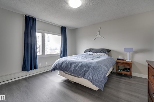 This room features gray flooring, light-colored walls, and a window with blue curtains - 1202 9909 110 Street Nw, Edmonton, AB - Indoor Photo Showing Bedroom