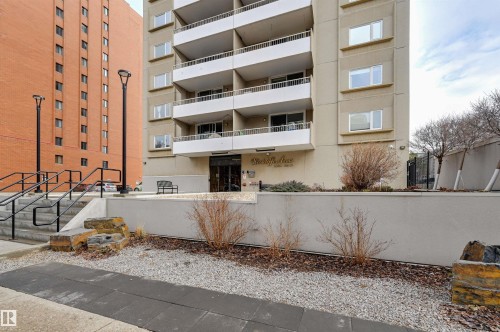 The property features a multi-story building with balconies, an entrance with a canopy, and landscaping with gravel and shrubs - 1202 9909 110 Street Nw, Edmonton, AB - Outdoor With Balcony