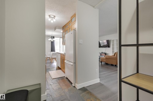 The property features a kitchen with wood cabinetry and a white refrigerator, and a living area with dark flooring - 1202 9909 110 Street Nw, Edmonton, AB - Indoor