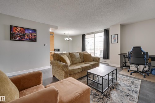 Living area featuring dark flooring, large windows with dark curtains, and light-colored walls - 1202 9909 110 Street Nw, Edmonton, AB - Indoor Photo Showing Living Room
