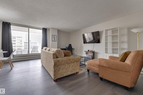Living area featuring dark flooring, white walls, and an expansive window with dark curtains providing views - 1202 9909 110 Street Nw, Edmonton, AB - Indoor Photo Showing Living Room