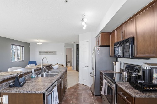 The kitchen features an island with a double basin sink, ample cabinetry, stainless steel appliances, and tile flooring - 115 6925 199 Street, Edmonton, AB - Indoor Photo Showing Kitchen With Double Sink