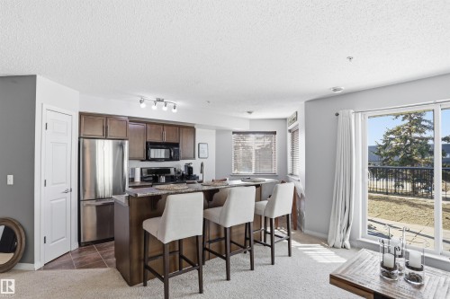 The open-plan living area features a kitchen with stainless steel appliances, dark wood cabinetry, and a granite countertop island with seating - 115 6925 199 Street, Edmonton, AB - Indoor