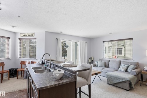 The open-concept living area features a kitchen island with a double sink and breakfast bar seating, carpeted flooring in the living room, and a sliding glass door providing external access - 115 6925 199 Street, Edmonton, AB - Indoor