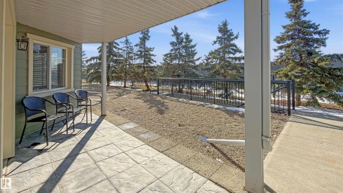 The property features a covered patio with stone-look pavers, offering an outdoor space with views of the surrounding landscape and evergreen trees - 115 6925 199 Street, Edmonton, AB - Outdoor With Deck Patio Veranda With Exterior