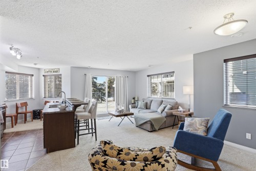 The bright living area features carpeted floors, light-colored walls, and large windows with blinds - 115 6925 199 Street, Edmonton, AB - Indoor Photo Showing Living Room