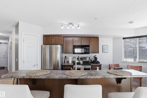 The kitchen features dark wood cabinetry, a stainless steel refrigerator, and a kitchen island with a stone-look countertop - 115 6925 199 Street, Edmonton, AB - Indoor Photo Showing Other Room
