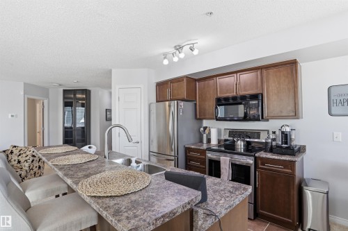 The kitchen features a breakfast bar with seating, a stainless steel sink with a high-arc faucet, and dark wood cabinetry - 115 6925 199 Street, Edmonton, AB - Indoor Photo Showing Kitchen With Double Sink