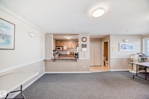 The interior features carpeted flooring, a kitchen area with wood cabinetry and a pass-through counter, and a restroom with a toilet and sink visible through an open doorway - 112 17150 94A Avenue, Edmonton, AB - Indoor Photo Showing Other Room