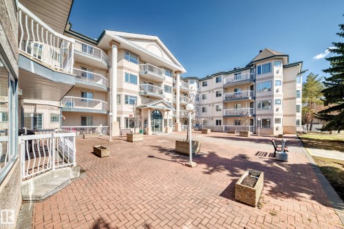 The property features a multi-story building with a light-colored exterior and multiple balconies, set on a brick paved courtyard - 112 17150 94A Avenue, Edmonton, AB - Outdoor