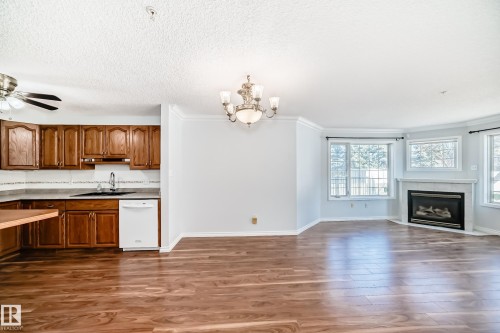 Open concept living area with wood flooring, a corner fireplace, and a kitchen featuring wood cabinetry, a white dishwasher, and a ceiling fan - 112 17150 94A Avenue, Edmonton, AB - Indoor With Fireplace