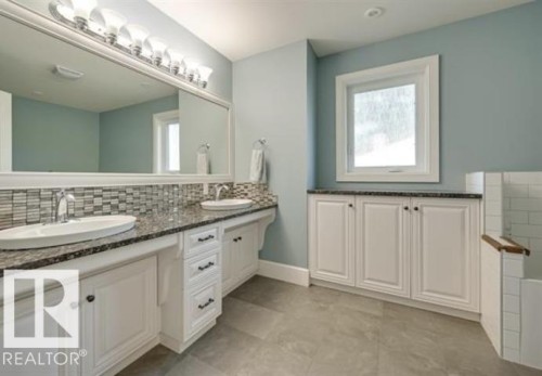 Bathroom featuring dual sinks with granite countertops and a tiled backsplash, white cabinetry for storage, and a window providing natural light - 10836 130 Street, Edmonton, AB - Indoor Photo Showing Bathroom
