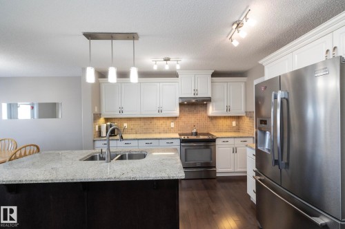 The kitchen features white cabinetry, stainless steel appliances, a tiled backsplash, and a central island with a sink and granite countertop - 132 Kirpatrick Way, Leduc, AB - Indoor Photo Showing Kitchen With Double Sink With Upgraded Kitchen