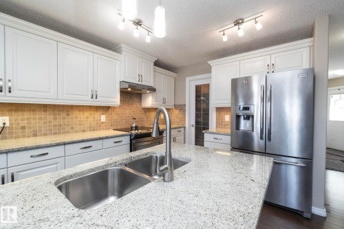 The kitchen features white cabinetry, a light-colored speckled countertop, a stainless steel refrigerator, and a stainless steel double basin sink with a contemporary faucet - 132 Kirpatrick Way, Leduc, AB - Indoor Photo Showing Kitchen With Double Sink With Upgraded Kitchen