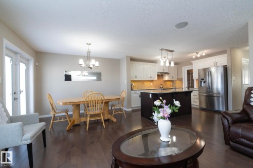 This open-concept living space features dark hardwood flooring, a dining area with a chandelier, and a kitchen with white cabinetry and a stainless steel refrigerator - 132 Kirpatrick Way, Leduc, AB - Indoor
