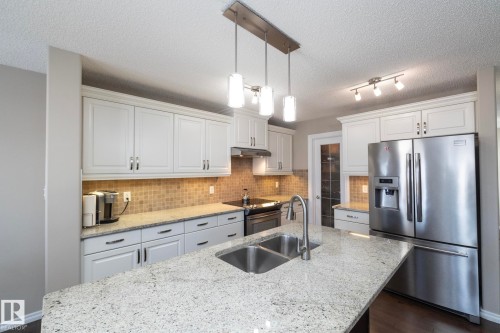 A modern kitchen featuring white cabinetry, a tiled backsplash, stainless steel appliances, and an island with a double sink and granite countertops - 132 Kirpatrick Way, Leduc, AB - Indoor Photo Showing Kitchen With Double Sink With Upgraded Kitchen