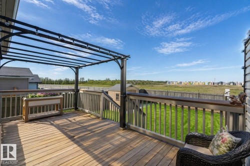 The property features an outdoor wooden deck with a dark-framed pergola, offering views of a grassy area and distant buildings under a clear sky - 132 Kirpatrick Way, Leduc, AB - Outdoor With Deck Patio Veranda With View With Exterior
