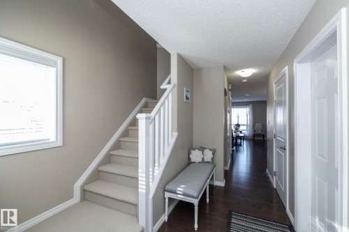 The property features a staircase with white railings and carpeted steps, a window with blinds providing natural light, and a hallway with dark wood flooring leading to a sunlit area - 132 Kirpatrick Way, Leduc, AB - Indoor Photo Showing Other Room