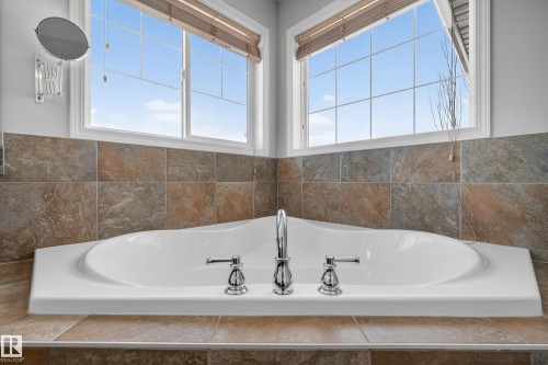 Corner bathtub with chrome fixtures, surrounded by large format tiles, and framed by two windows with blinds - 20714 56A Avenue, Edmonton, AB - Indoor Photo Showing Bathroom