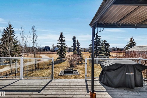 The property features an outdoor deck with wooden planks and a glass railing, providing views of a landscaped area with mature trees and a paved pathway - 5201 40 Avenue, Beaumont, AB - Outdoor