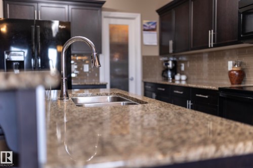 The kitchen features dark cabinetry, granite countertops, a stainless steel sink with a pull-down faucet, and a tiled backsplash - 130 Kirpatrick Way, Leduc, AB - Indoor Photo Showing Kitchen With Double Sink