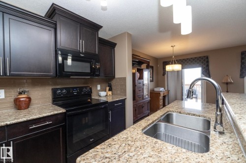 The kitchen features dark wood cabinetry, granite countertops, a stainless steel double sink with a gooseneck faucet, and a black range with an overhead microwave - 130 Kirpatrick Way, Leduc, AB - Indoor Photo Showing Kitchen With Double Sink